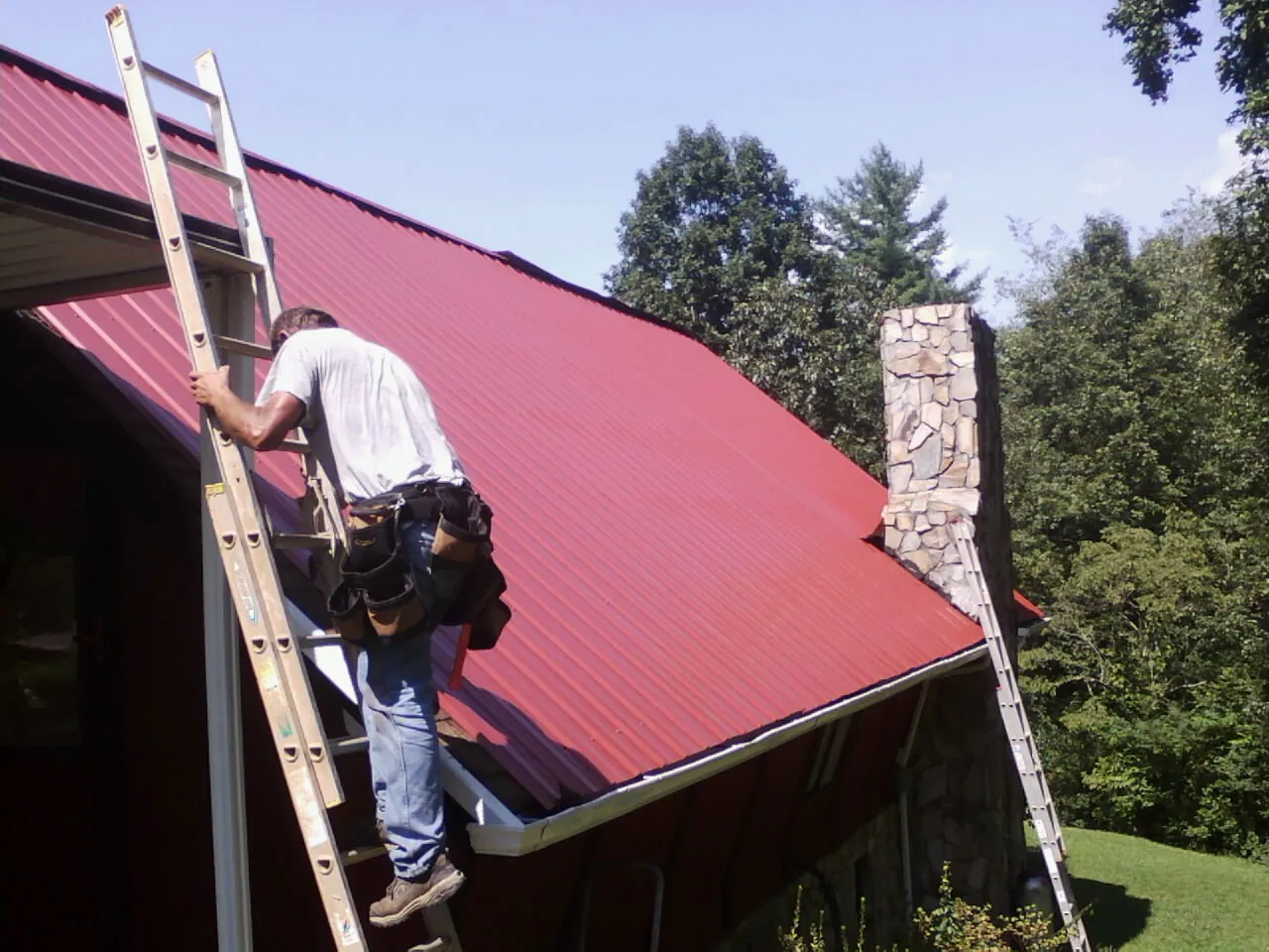 Roofer on a ladder beside a red metal roof and stone chimney — Soffit & Fascia Repair by Roofing Brothers in Emden